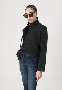 Pulover tricotat de culoare gri închis, cu guler înalt, manșete bleonate și o croială lejeră, asortat cu blugi și ochelari de soare geometrici negri.
