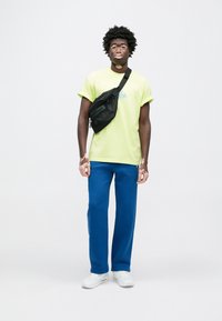 Neónovo žlté tričko s modrou grafikou, skombinované s modrými teplákmi a bielymi teniskami. Čierna crossbody taška je nesená cez jedno rameno.