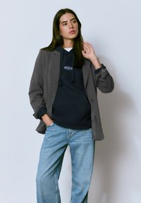 Blazer gris sobre una sudadera azul marino con cordones, combinado con jeans de color azul claro. Presenta un ajuste casual y detalles de costura visibles.