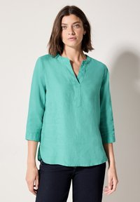 Camicia di lino turchese con scollo a V, maniche a tre quarti e vestibilità rilassata; presenta un orlo leggermente curvato e tessuto testurizzato.