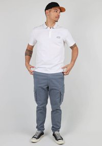 Chemise polo blanche avec un col, pantalon cargo gris avec plusieurs poches et baskets noires avec semelles blanches, sur un fond uni.