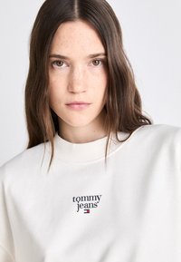 Svetlo béžová mikina so zaobleným výstrihom, ribkovanými manžetami a centrálnym logom v čiernej a červenej farbe s nápisom "tommy jeans."