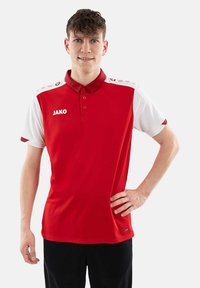 Polo rosso con maniche corte bianche, dotato di colletto, tre bottoni e logo "JAKO" sul petto. Tessuto morbido, vestibilità atletica.