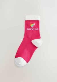 Lys pink sok med hvid hæl, tå og manchet, der har et regnbuehjerte og ordet "SONNTAG" i hvid tekst.