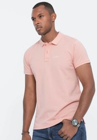 Vaaleanpunainen puuvillapolo paita, jossa on klassinen kaulus, lyhyet hihat ja kolme nappia, ja joka sisältää hillityt ommel yksityiskohdat rinnassa.