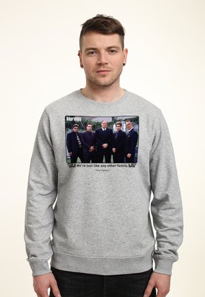 Mężczyzna w szarej bluzie z nadrukiem zdjęcia pięciu mężczyzn w garniturach i napisem „Jesteśmy jak każda inna rodzina” z serialu The Sopranos.