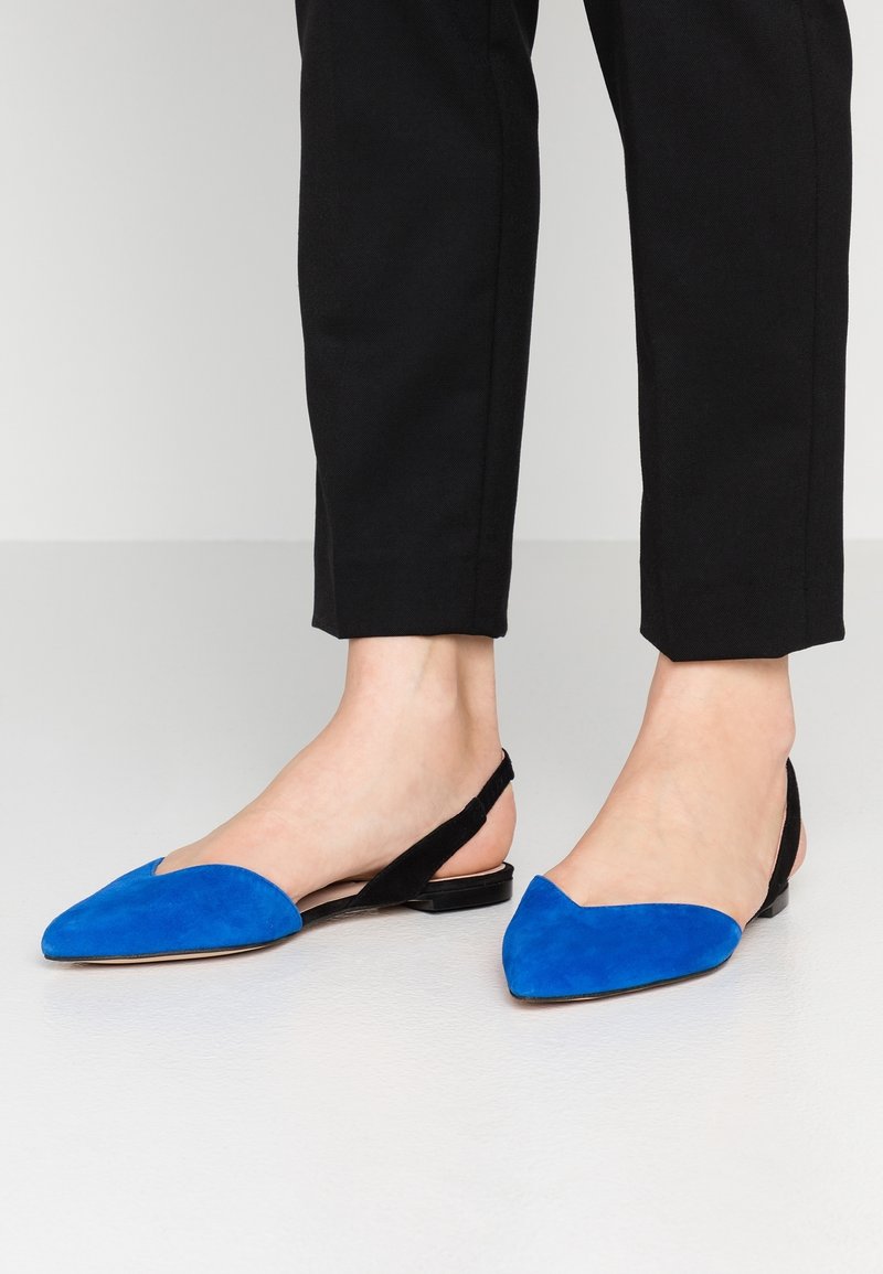Femme portant un pantalon noir raccourci avec des ballerines bleu et noir à bout pointu et bride arrière, sur un sol pâle.
