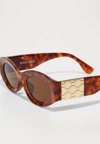 Tortoiseshell sunglasses with brown lenses, bold oval frame, and a gold hexagonal accent detail on the temples. Brand name visible on side.