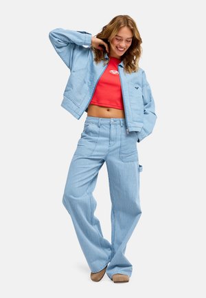 Tânără zâmbitoare, purtând o jachetă și pantaloni cu dungi albastre deschise și un top roșu scurt, stând cu o mână în spatele capului.