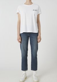 Bílé bavlněné tričko s krátkými rukávy a kapsou s květinovým motivem, v kombinaci s modrými džínami s rovnými nohavicemi a bílými teniskami.