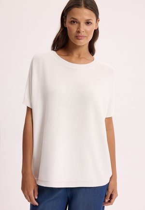 Kobieta o ciemnych włosach, ubrana w luźną białą bluzkę z krótkim rękawem i niebieskie spodnie, stojąca na jasnym tle.