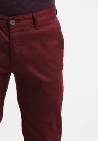 Pantaloni di cotone color bordeaux con taglio dritto, dotati di chiusura con bottone e tasche laterali. Texture liscia con dettagli di cuciture minime.