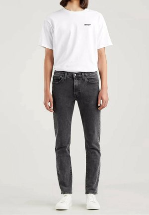 Person trägt weißes Levi's T-Shirt, schwarze Slim-Fit-Jeans und weiße Sneakers und steht vor einem neutralen Hintergrund.