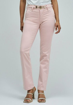 Mujer con pantalones rectos color rosa claro con cinturón de cadena dorada y sandalias beige de tacón alto con tiras sobre fondo liso.