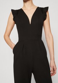 Svart jumpsuit med djup V-ringning, volangärmar, spetsdetaljer vid axlarna och sidofickor; strukturerad tyg med en jämn textur.