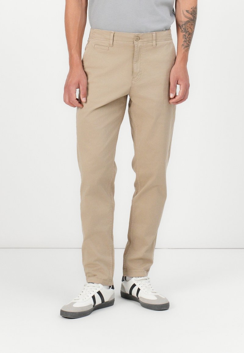 Homme portant un pantalon beige droit, des baskets blanches avec des rayures noires et une chemise gris clair, debout devant un fond blanc uni.
