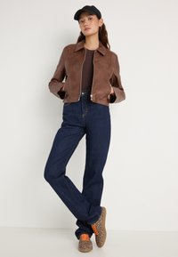 Jaqueta curta de couro marrom com colarinho, jeans de cintura alta azul escuro e ténis com bolinhas e atacadores laranja. Boné simples e brincos de argola.