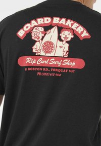 Twee cartoonfiguren die een surfplank vasthouden met de tekst "Rip Curl Surf Shop" en contactgegevens op een zwart t-shirt.