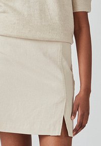 Falda beige clara hecha de material suave, con corte recto, aberturas laterales y un detalle de costura mínima. La modelo la sostiene.