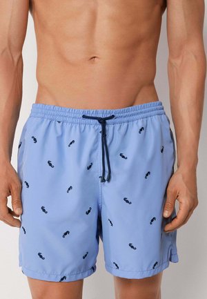 Mann trägt hellblaue Badeshorts mit kleinen dunklen Seepferdchenmustern und schwarzem Kordelzug, gezeigt vom Oberkörper bis zur Mitte des Oberschenkels.