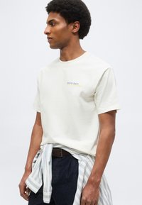 Άνδρας που φοράει εκρού μπλουζάκι με μικρό κείμενο "Oyster Party", σκουρόχρωμο παντελόνι και ριγέ πουκάμισο δεμένο στη μέση, κοιτάζοντας αριστερά.