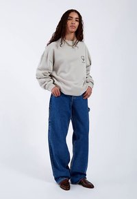 Světle šedá volná mikina s logem srdce, doplněná volnými modrými džínami. Model stojí s rukama v bok, na nohou má hnědé boty.