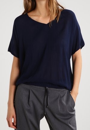 Kobieta ubrana w luźną granatową bluzkę z dekoltem w serek, wpuszczoną w szare spodnie z sznurkiem, z rękami w kieszeniach, na jednolitym tle.