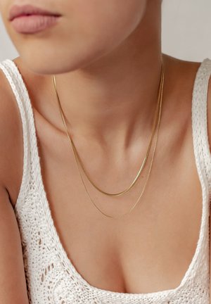 Femme portant deux délicats colliers en chaîne dorée sur un débardeur blanc en maille, montrant le cou, les épaules et une partie du visage.