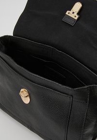Schwarze Lederhandtasche mit strukturiertem Oberflächen, goldenen Beschlägen und einer Vorderklappe zum Öffnen. Das Innere ist mit schwarzem Stoff ausgekleidet.
