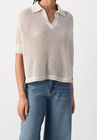 Jasnobeżowy dziany sweter polo z dekoltem w serek, krótkimi rękawami i ażurową teksturą. Nosi się go na niebieskie spodnie dżinsowe.