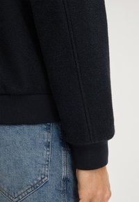 Zwarte fleecetruwe met textuuroverzicht, geribde manchetten en een getailleerde zoom, gecombineerd met lichtblauwe spijkerbroeken. Close-up van de mouw en hand.