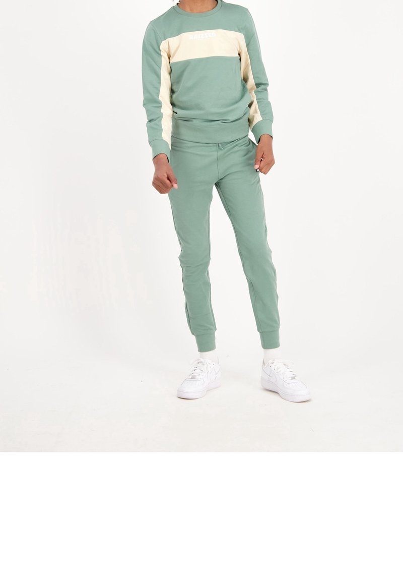 Sudadera verde y pantalones jogger a juego, hechos de suave algodón. La sudadera presenta una franja crema en el pecho con texto. Zapatillas blancas.