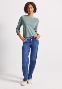 Vrouw staat met haar handen in haar zakken en draagt een salie groene lange mouwen top, blauwe high-waisted jeans en witte sneakers tegen een witte achtergrond.
