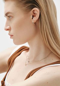 Pendiente de aro dorado con acento de turquesa, cadenas de plata en capas con un pequeño colgante, usado por un modelo con largo cabello rubio liso.