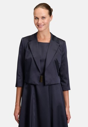 Boléro en satin marine à manches trois-quarts et à coupe courte, porté par-dessus une robe assortie. Il présente une texture lisse et un col structuré.