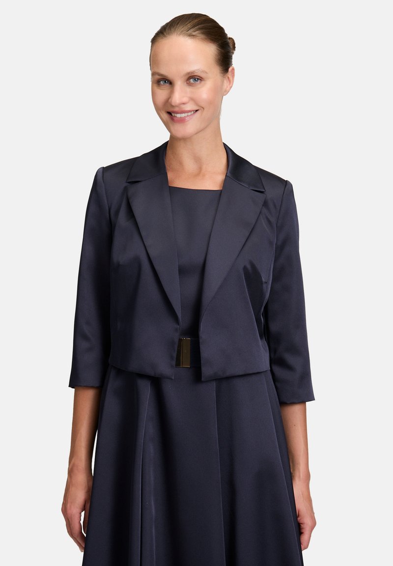 Granatowa satynowa bolerko z rękawami o długości trzech ćwiartek i skróconym kroju, noszona na dopasowanej sukience. Ma gładką fakturę i usztywniony kołnierz.