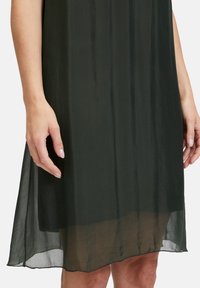 Schwarzes, durchscheinendes Kleid mit einer weichen, fließenden Textur. Verfügt über eine geschmeidige Silhouette und einen Saum, der sanft die Hand berührt, die sich in seiner Nähe befindet.