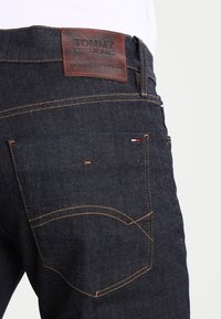 Mörk denimjeans med en brun läderlapp märkt "TOMMY JEANS." Har orange sömmar och två bakfickor med böjda sömmar.