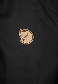 Juodas audinys su mažu šviesiai rudos odos lapės logotipu, turinčiu lygų paviršių, prieš subtilų austą foną.
