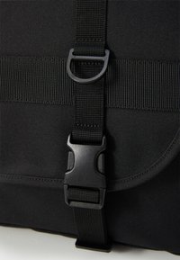 Sac en tissu noir avec une boucle en plastique et une sangle en toile. Texture lisse avec une forme plate et rectangulaire et un détail de rabat incurvé.