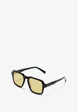 Vierkante zonnebril met een dik zwart montuur, geel getinte glazen en groen gemêleerde accenten op de pootjes. Heeft een glanzende afwerking.