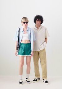 Chemise à manches courtes bleu clair, crop top blanc, shorts verts avec dentelle, lunettes de soleil noires ; chemise à manches courtes beige, pantalon kaki, baskets.