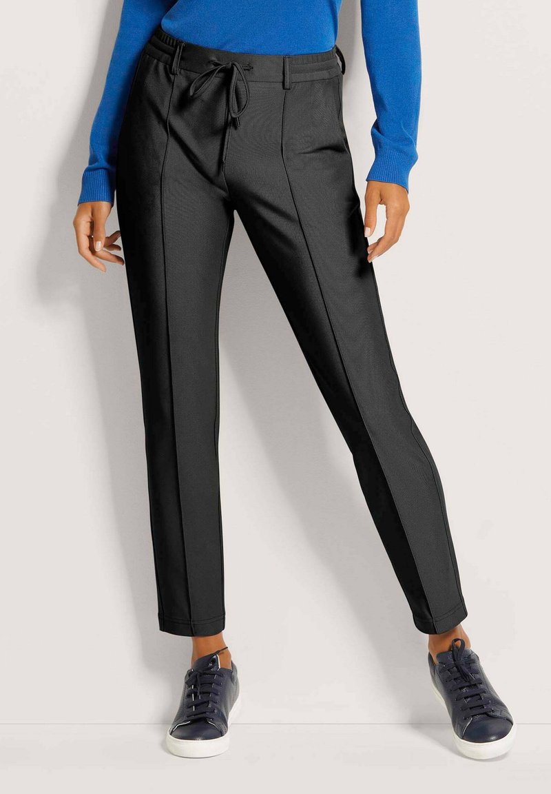 Vrouw in zwart op maat gemaakte broek met een trekkoord taille, een blauwe lange mouwen top en zwarte sneakers met witte zolen.