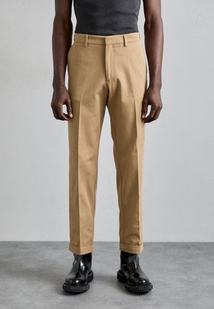 Beige chinos egyenes szabással, középmagas derekú derékkal és elől hajtással, fekete fényes bokacsizmával párosítva. Simaságot biztosító pamut anyag.
