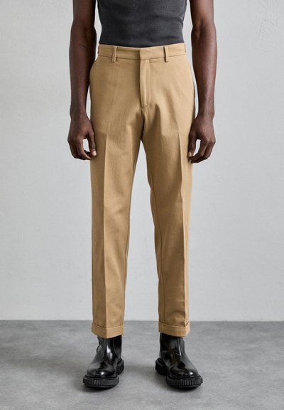 Chinos beige à coupe droite, taille mi-haute, avec pli avant, associés à des bottines noires brillantes. Matériau en coton lisse.
