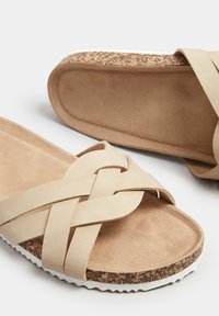 Sandales à brides croisées beige avec un dessus doux en suède, un semelle intérieure en liège texturé, une semelle en caoutchouc blanche et un design plat.