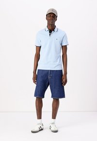 Lysblå polo skjorte med sort krave og trim, parret med mørke denim shorts. Modellen bærer en grå kasket og hvide sneakers.