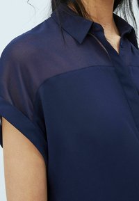 Mörkblå blus med korta uppvikta ärmar, krage med knappar och en genomskinlig övre panel, med en mjuk textur och skräddarsydd passform.