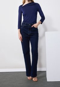 Vrouw in een marineblauwe asymmetrische langslijf top, donkerblauwe high-waisted jeans en gouden sandalen met open tenen staat naast een witte zuil.