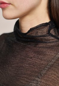 Schwarzer, transparenter Rollkragenpullover aus strukturiertem Stoff, mit feinen Punktmustern und zarter Borte am Kragen, die ein leichtes Design hervorhebt.